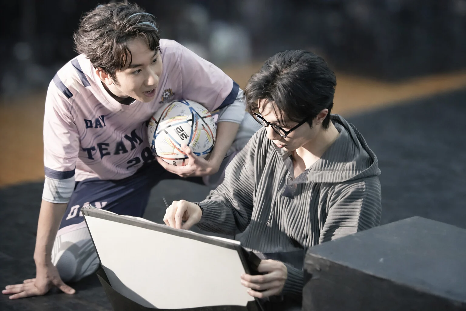 Reincarnated as a Convenience Store: a performer in a football kit holding a ball leans towards another reading from a sketchbook