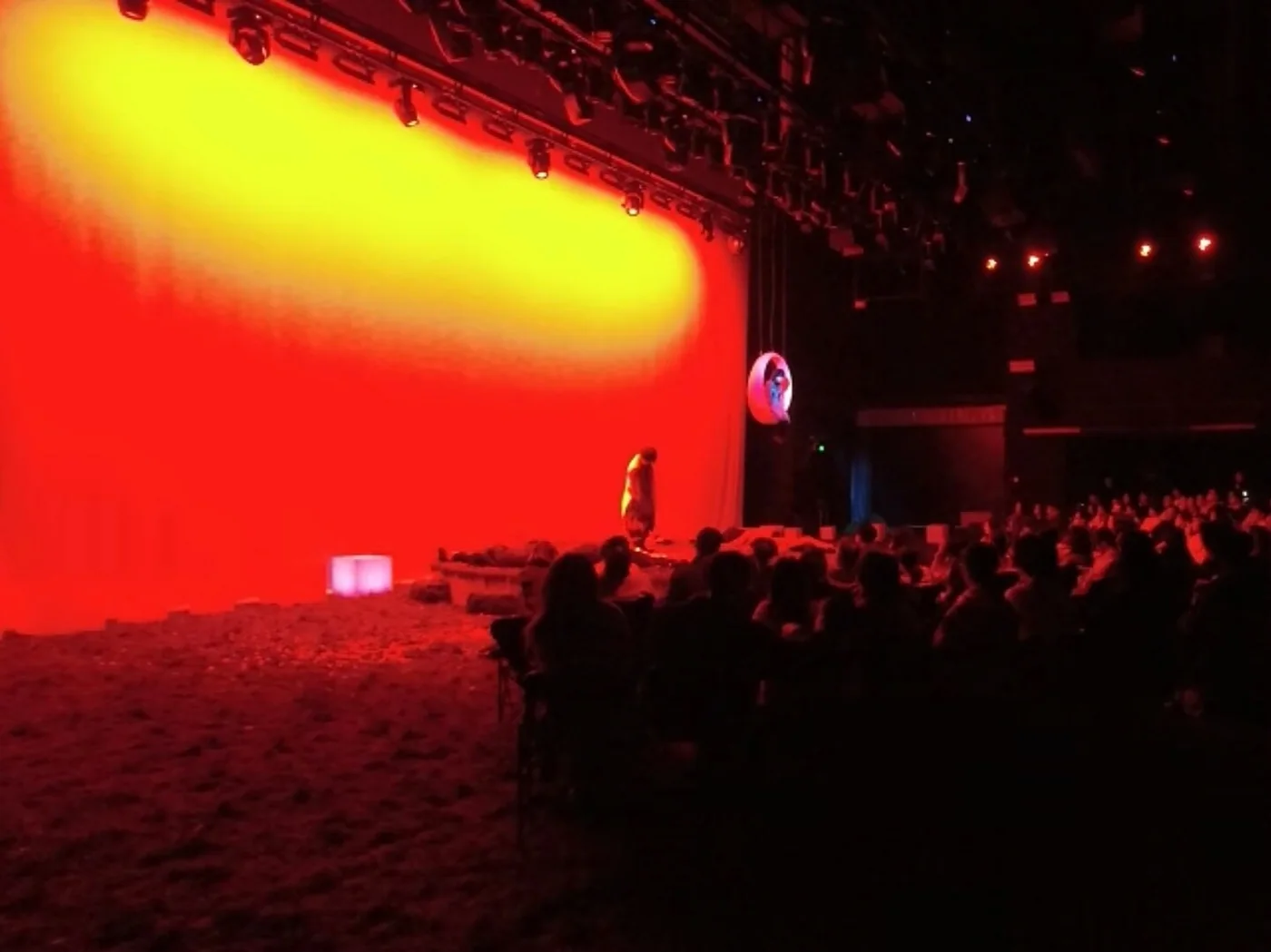 The Moon, The Moon: a performer before a red and yellow lit backdrop, with the audience in the foreground