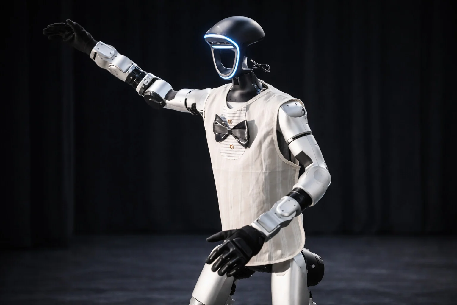 Reincarnated as a Convenience Store: a Unitree humanoid robot in a white bib and bow tie, one arm raised, against a dark stage